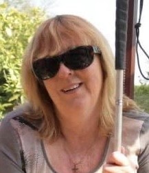 Headshot of Lynne Rennison, Lancashire SLC member. Lynne is sitting outside, wearing dark sunglasses. She is holding her white cane up in her hand.
