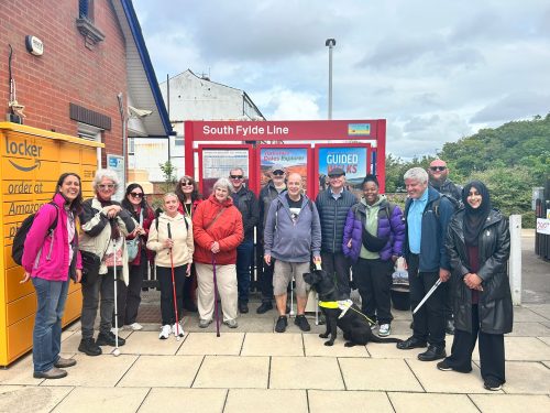 A group of seven people of mixed ages, genders and ethnicities. Some are holding white canes and there is one person with a guide dog. The group is standing in front of a red sign that reads, 'South Fylde Line' at a train station. There is an Amazon locker to the left and trees in the background.