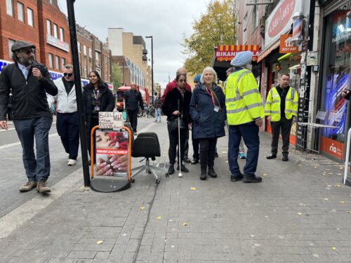 Sight Loss Council volunteers and staff stand on a busy pavement in front of shops with Hounslow Council representatives, two who are wearing high-visibility jackets. A freestanding advertising board is on the pavement and an office chair obstruct part of the walkway.