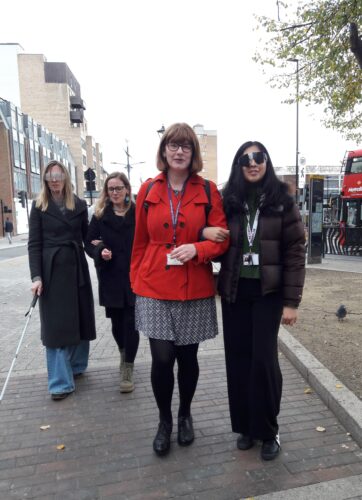 Four people walking along a pedestrian area in Hounslow Town Centre. Councillor Dunne is on the left of the front row wearing a bright red coat and is guiding another female council representative wearing sim specs by linking arms. Another person behind them being guided wears sim specs and uses a white cane. Buildings, a bus stop, and a red double-decker bus are visible in the background.