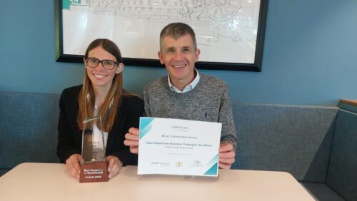 Sight Loss Council (SLC) Coordinator Lauren (left) and SLC Engagement Manager Dave Smith (right) are seated at a table. Lauren is holding a glass award with a wooden base that reads 'Bus Centre of Excellence Award, 2025'. The glass section is engraved with 'Winner of the Collaboration Award'. Dave is holding a certificate titled 'Certificate of Achievement', which recognises the BCoE Collaboration Award for Vision Impairment Awareness Training for Bus Drivers, presented by Thomas Pocklington Trust. The certificate features the CIHT logo, a signature, and an awards emblem.