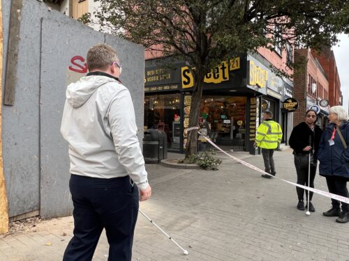 A Hounslow Council representative wearing a light grey jacket, sim specs and holding a white cane approaches a taped-off area on a pavement which is blocking his path. Behind the tape, there is a tree and a shop called “Street 21” with a yellow sign. Another person in a high-visibility jacket stands nearby, alongside a Sight Loss Council volunteer and female Council representative.