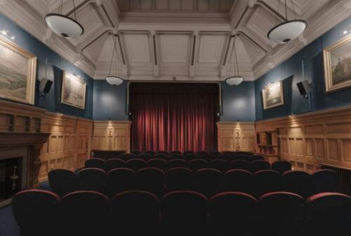 Interior of Guildhall Cinema showing rows of red and black seats facing a stage with closed red curtains. The room has wood-panelled walls, blue upper walls, framed paintings, and a decorative white coffered ceiling with hanging lights.