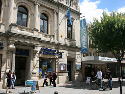 Exterior view of Guildhall building in Gloucester with stone façade, arched windows, and a blue vertical banner reading “Guildhall.” People are walking and cycling along the pavement. A Cheltenham & Gloucester bank branch is visible at street level.