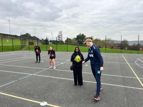 A young female participant is stood smiling in an outdoor sports court holding a yellow rugby ball while another individual in sportswear provides guidance. Two more people stand further back, and the court is marked with white and blue lines. A grassy slope and fencing are in the background.
