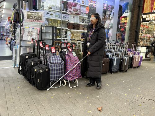 Zahra Ali, Senior Strategic Transport Planner at Hounslow Council holds a white cane and wears sim specs. She is walking in front of a shop window displaying luggage and household items. Multiple suitcases and shopping trolleys are lined up on the pavement outside the store, causing an obstruction.