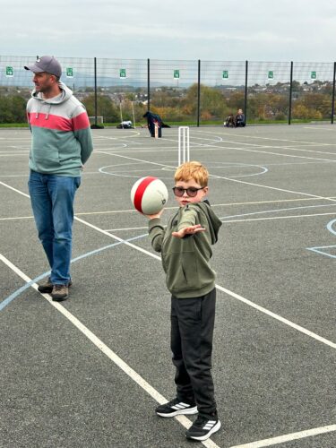 A child stands on an outdoor sports court holding a red-and-white ball in one hand, with the other arm extended as if preparing to throw. The child is wearing a green hoodie, black pants, and sneakers. Behind the child, an adult in a grey and red hoodie and jeans stands on the court. The background shows a fenced area with markings for multiple games and a few people seated near the fence. The sky is overcast.