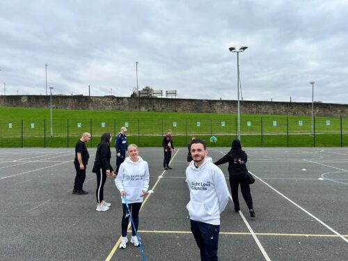 Sight Loss Council Engagement Manager, Kelly Barton (left), and SLC Co-ordinator, David Parfett (right), are stood in the foreground wearing white hoodies with the SLC logo printed on them. They are smiling at the camera. Kelly is holding a blue navigation cane. In the background, six event participants are spaced out on an outdoor sports court, throwing a ball to one another that is visible mid-air. The balll is specially adapted and has a bell inside that makes a noise. A grassy embankment and fence line the background.