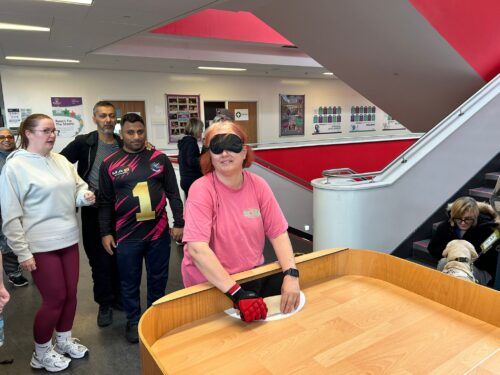 A participant wearing gloves and a blindfold stands at a wooden showdown table, ready to play. Several others are gathered around faced towards the showdown table. They are pictured in an indoor area of the school, which has red and cream walls, stairs, and posters on the walls.