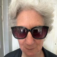 Headshot of Steph Syson. She is wearing dark glasses, has grey wavey hair and a black top. She is smiling at the camera.