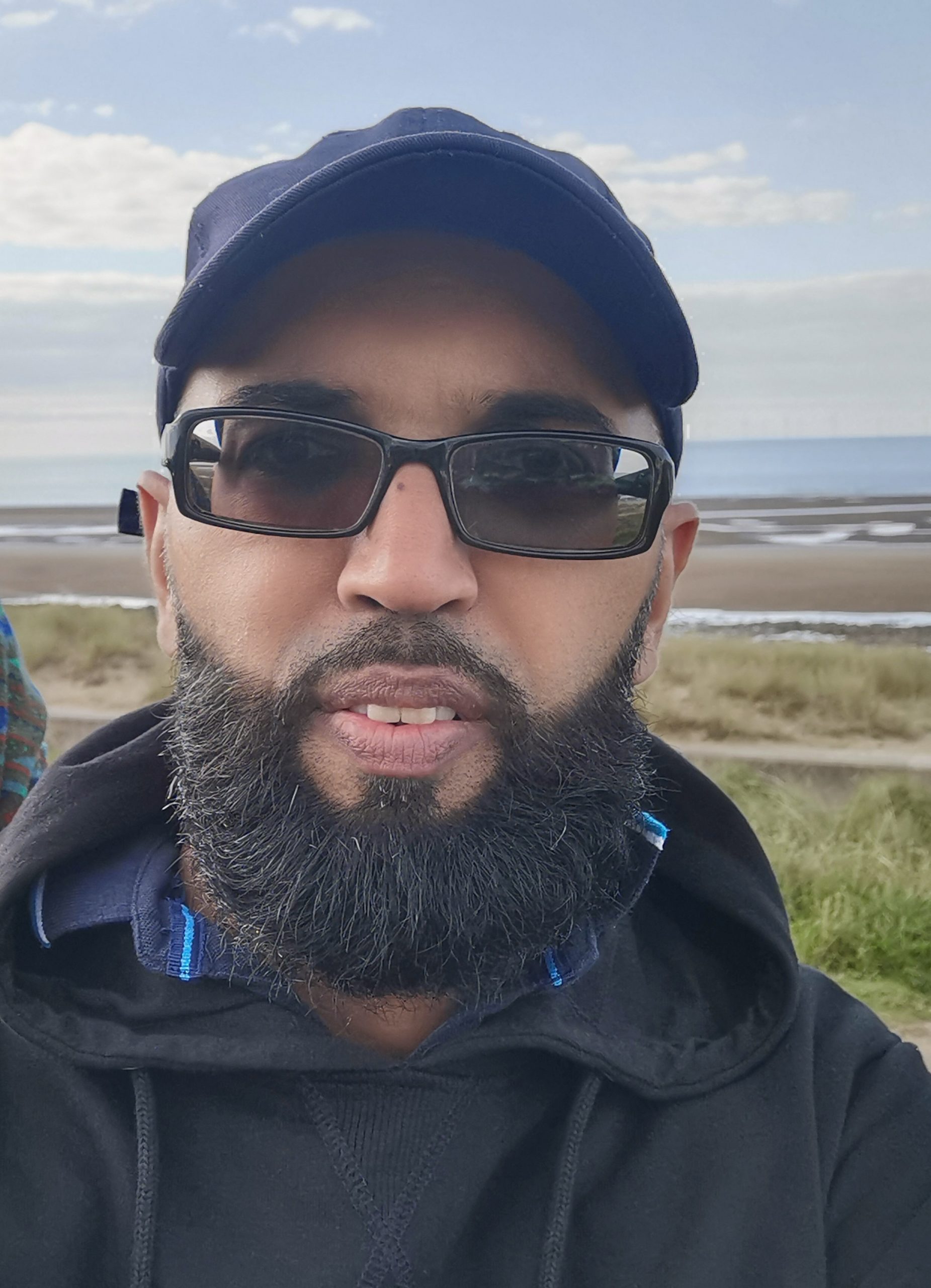 Photo of Amin, Greater Manchester SLC volunteer. Amin is outdoors in a countryside setting. He is wearing a dark hat and dark sunglasses. Amin has a dark beard, and is smiling at the camera.
