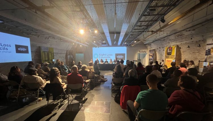 Image taken from behind the audience, looking forward to the panel of guests speakers. There is a large white screen behind the speakers which says: Sight Loss Councils, with a small black box to the side which says "RIP Her Majesty."