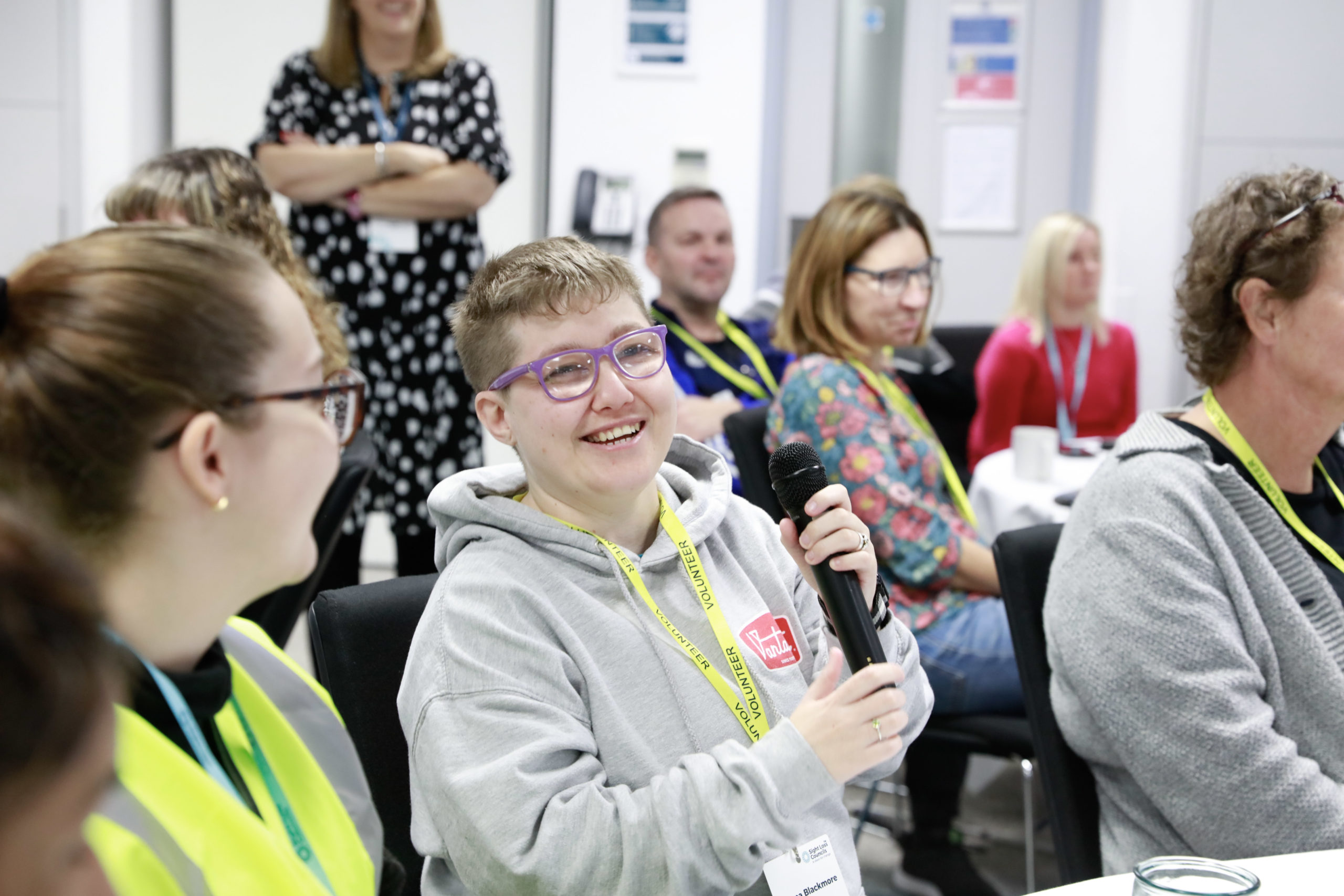 Sight Loss Council volunteer Emma holds a microphone ready to share her views at the Sight Loss Council volunteer conference 2022. She is in a room with other conference delegates.