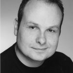 Black and white headshot of Liam O'Carroll. His head is turned slightly to the side and he is smiling.