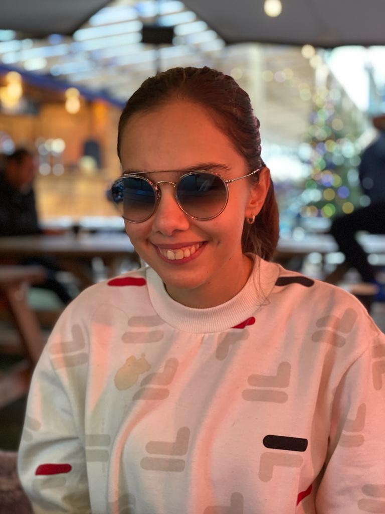 Image shows Zehra, Student Voices member, smiling at the camera. Her hair is pulled back in a ponytail, she is wearing a white jumper, and sunglasses.