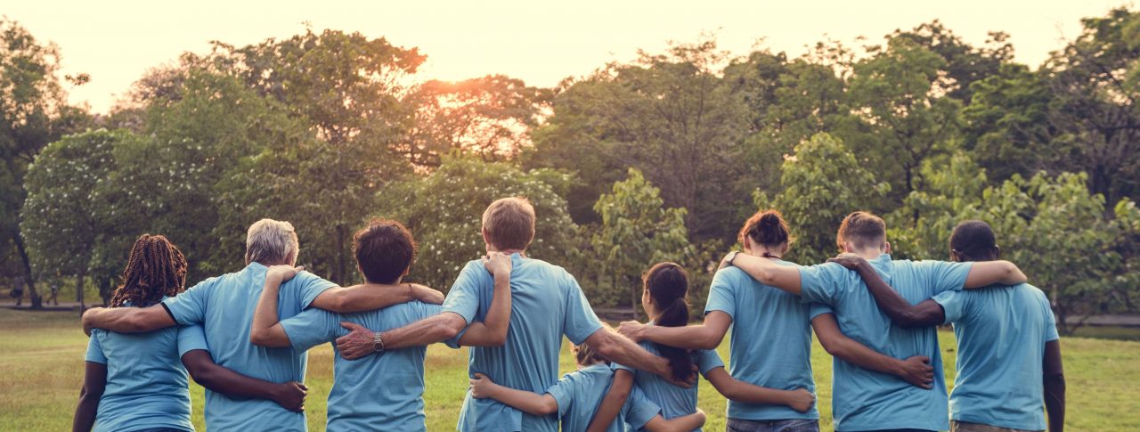 Group of volunteers with their arms around each other, facing away from the camera. They are outside and are all wearing light blue t-shirts.