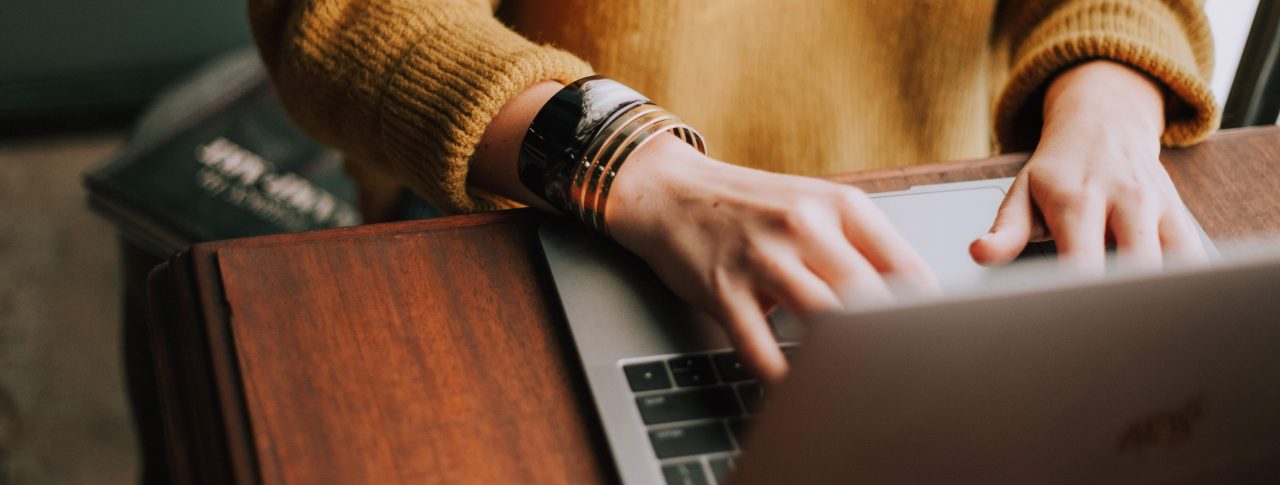 Close up of a woman's hands, typing on a silver laptop. She is wearing a mustard yellow jumper, and has two bangles on her wrist.