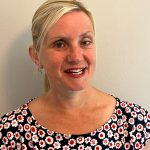 headshot of Kelly Barton, Engagement Manager for the north west. Kelly is standing against a white wall and smiling at the camera. Her hair is pulled back into a pony tail. She is wearing a black top with red and white flowers on it.