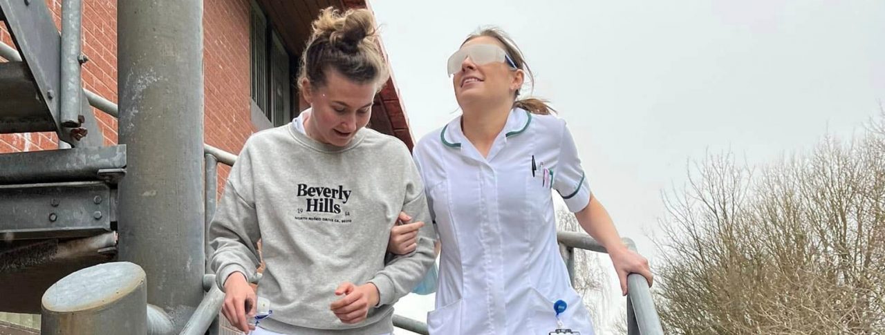 Two nurses from Southport Hospital, walking down a staircase as part of a vision awareness session. One nurse is being guided, whilst wearing simulation spectacles.