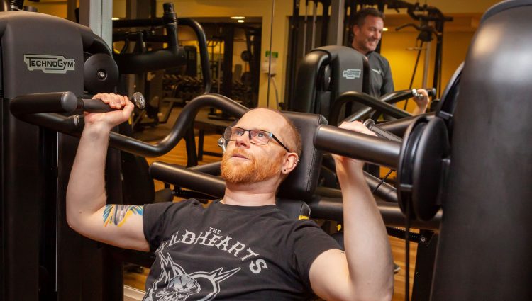 A partially sighted male, using weights in the gym. An instructor is seen in the background, smiling encouragingly.
