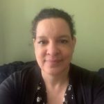 Denise, London SLC member, is seated, facing the camera, and smiling. Her hair is tied back.