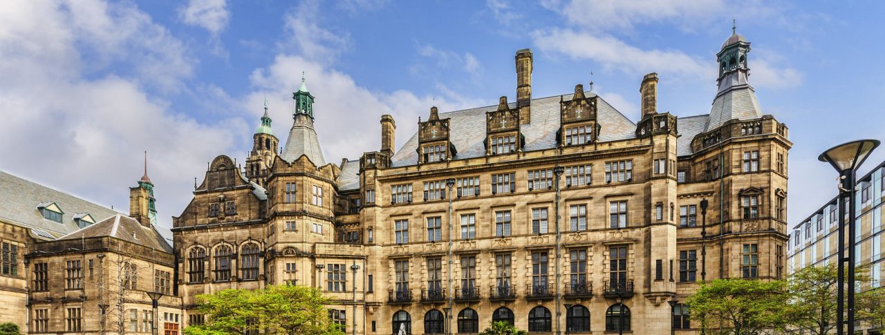Landscape image of Sheffield Town Hall, a Grade 1 listed building built with Stoke stone and ornate carvings. In front of the town hall is a circular fountain, grass and trees.