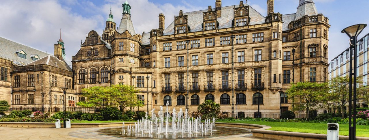 Landscape image of Sheffield Town Hall, a Grade 1 listed building built with Stoke stone and ornate carvings. In front of the town hall is a circular fountain, grass and trees.