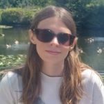 Lauren Eade headshot. Lauren has long brown hair and is wearing sunglasses and a white t-shirt sitting in a garden in the sun.