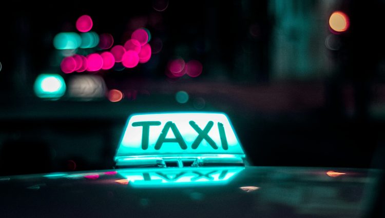 Close up of a taxi light, lit up in blue and white. The background is dark - night time - with some twinkling lights shown in the background.