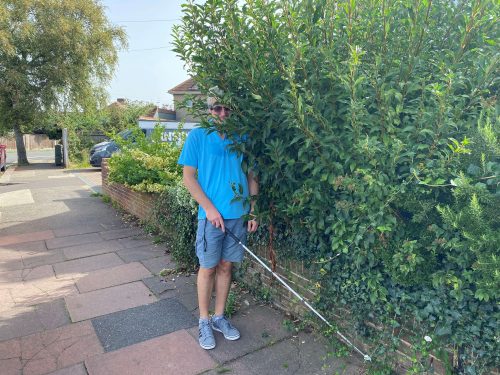 Sussex SLC member, Clinton walking with his long cane, into a large garden hedge which is protruding onto the pavement.