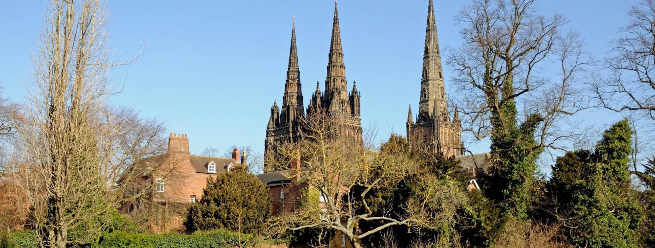 Landscape image of Lichfield Cathedral in Staffordshire. The photo is taken from the other side of the river. Two people are sat on a wooden park bench in front of the cathedral, and large trees frame the cathedral.