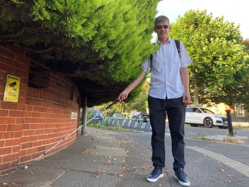 Dave Smith, Engagement Manager for South East England, is pictured next to a large hedge which is overhanging into the pavement. Dave is holding his long cane and looking at the camera.