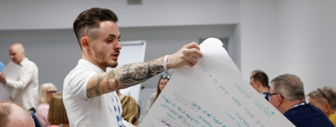 David Parfett, SLC Coordinator for the North West, reading from a large sheet of paper with notes on it at a table of delegates during a session at the SLC conference.