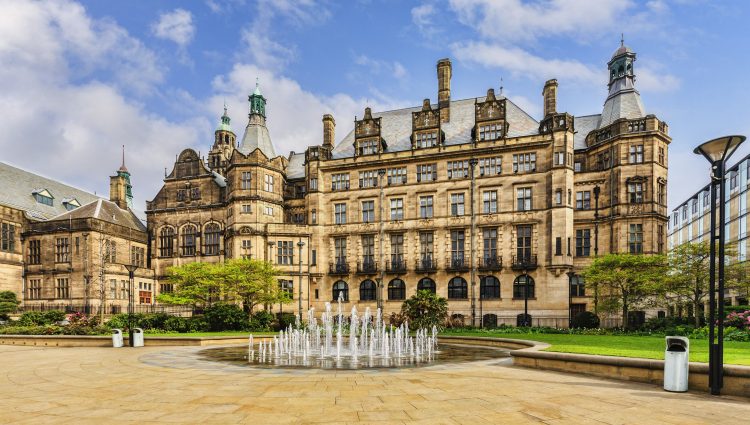 Landscape image of Sheffield Town Hall, a Grade 1 listed building built with Stoke stone and ornate carvings. In front of the town hall is a circular fountain, grass and trees.