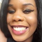 Headshot of Monique, North London SLC member. Monique has long dark hair and winged eyeliner in this photo. She is smiling broadly at the camera.