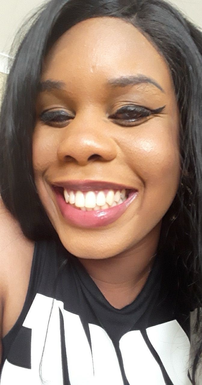 Headshot of Monique, North London SLC member. Monique has long dark hair and winged eyeliner in this photo. She is smiling broadly at the camera.