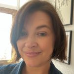 Headshot of Nicki Guy, Senior Engagement Manager for South England. She is standing in a hallway in front of prints on a wall, smiling at the camera.