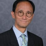 Paul Hsu, North London SLC member. Paul is wearing a dark suit and stood against a grey background. He is wearing glasses, and smiling at the camera.