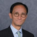 Paul Hsu, North London SLC member. Paul is wearing a dark suit and stood against a grey background. He is wearing glasses, and smiling at the camera.