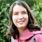Ellie Wallwork headshot. Ellie has brown hair and is wearing a pink hoodie while smiling at the camera with trees in the background.