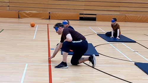 Four participants playing goalball. They are blind folded and sitting on mats in a sports hall. The participant closet to the camera is a man in black t-shirt and shorts, kneeling with his hand touching a red line on the floor.