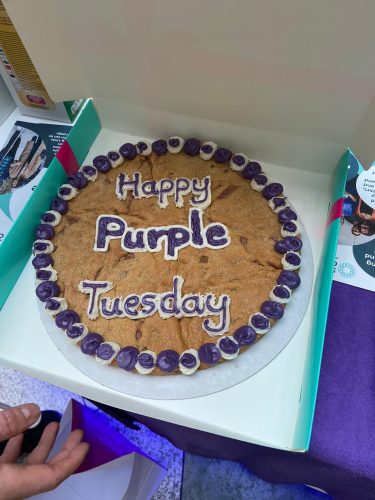 Image shows a large cookie with 'Happy Purple Tuesday' in purple and white icing in a cardboard box.