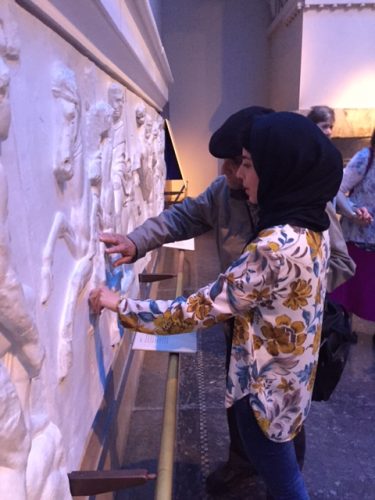 A woman wearing a floral shirt and hijab interacting with a tactile display on a wall featuring raised artwork of figures and animals.