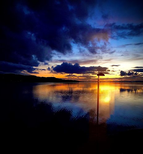 The image depicts a serene sunset scene and replicates the loss of peripheral vision from the left side. In the foreground, a silhouette of a streetlight is partially visible, casting a warm glow on the water's surface. The water is calm, reflecting the colours of the sky. The sky is filled with clouds that are illuminated by the setting sun, creating a mix of orange, pink, and purple hues. In the distance, a silhouette of land or an island can be seen, adding depth to the scene. The overall atmosphere is peaceful and tranquil, with the soft light of the sunset creating a beautiful and calming effect. 