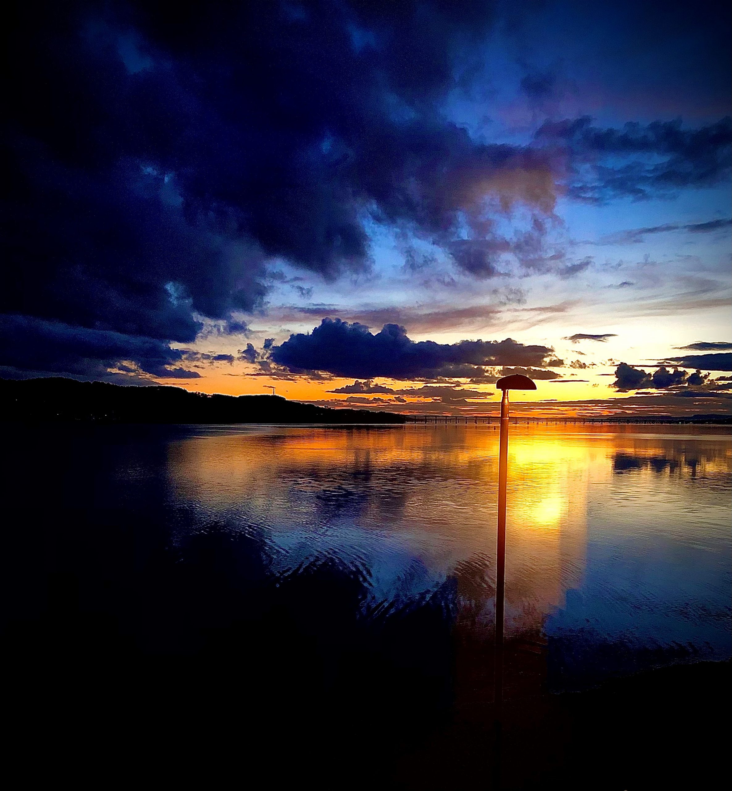 A striking painting of a sunset over a calm lake. The sky is a dramatic mix of deep blue clouds and warm orange, pink and yellow hues reflecting on the water. A single lamppost stands near the water’s edge, silhouetted against the sky.