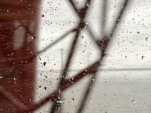 The image depicts raindrops on a glass window in sharp focus with a blurred background of a red metal rail bridge and a river in the distance. This highlights very short sightedness. The raindrops are scattered across the glass, and the background is indistinct, likely due to the rain and the movement of the camera. The raindrops vary in size, with some larger and more prominent, while others are smaller and more numerous. The raindrops appear to be falling from the top of the window, and the glass is wet from the rain. The raindrops are reflecting the light, creating a shimmering effect on the glass. 
