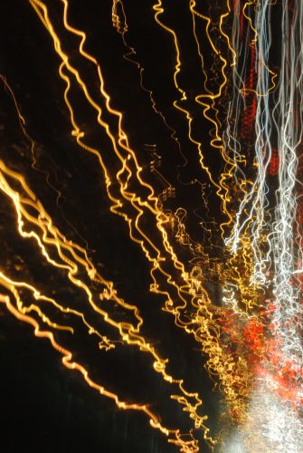 The image depicts a night scene with bright lines highlighting the patterns some people with Charles Bonnet Syndrome may see. It appears to be a long exposure photograph of a night-time road scene. It depicts various streaks of light that resemble trails of headlights from cars and other vehicles. The streaks of light are in different colours, predominantly yellow and white, with some red streaks as well. These streaks are elongated and wavy and start in a concentrated spot in the top left corner and dispersing down and outwards towards the right bottom corner. These appear to indicate motion over time. The background is dark, suggesting it is nighttime, with no other discernible objects or details visible in the scene. 