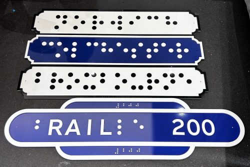 The tactile sign in a heritage style for train stations is located on the far wall. Two signs are located here. The first sign is heritage blue with a white boarder. The tactile element is braille and reads Colchester. The second sign is located underneath and is also in heritage blue with a white boarder and reads ‘b’ in braille, rail in tactile lettering and ‘le’ and the number ‘200’ in braille to link the 200 years of braille and the modern railway which celebrates 200 years this year. The numbers 200 are also in smaller braille font at the top and bottom of the sign.    