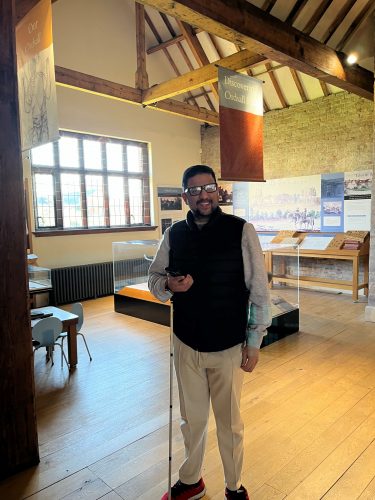 Greater Manchester SLC member Abu pictured smiling at the camera with his phone in his hand to access the audio descriptions at Ordsall Hall.