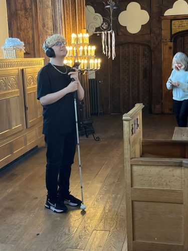 Greater Manchester SLC member Anthony Gough pictured standing with his cane in his hand and wearing headphones to access the in app audio descriptions at Ordsall Hall.